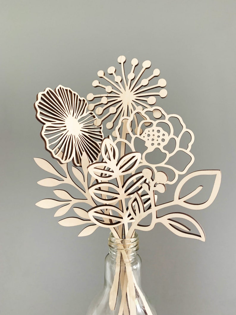 May include: A bouquet of wooden flowers in a clear glass bottle. The flowers are cut from light-colored wood and feature various floral and leaf designs. The arrangement includes a large, open flower, a spiky bloom, and several leafy stems.