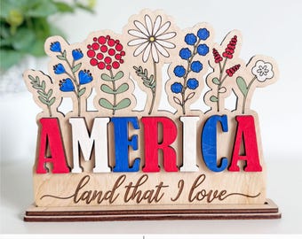 May include: A wooden sign with the word "AMERICA" in red, white, and blue letters, with the phrase "land that I love" below. The sign features a floral design on top and is laser-ready.