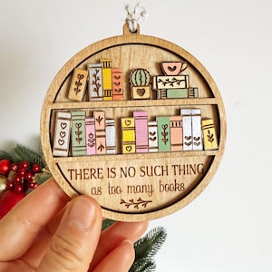 May include: Wooden, round ornament featuring a bookshelf design with colorful books and a cactus. The text "THERE IS NO SUCH THING as too many books" is engraved below the shelf. A decorative item for book lovers.