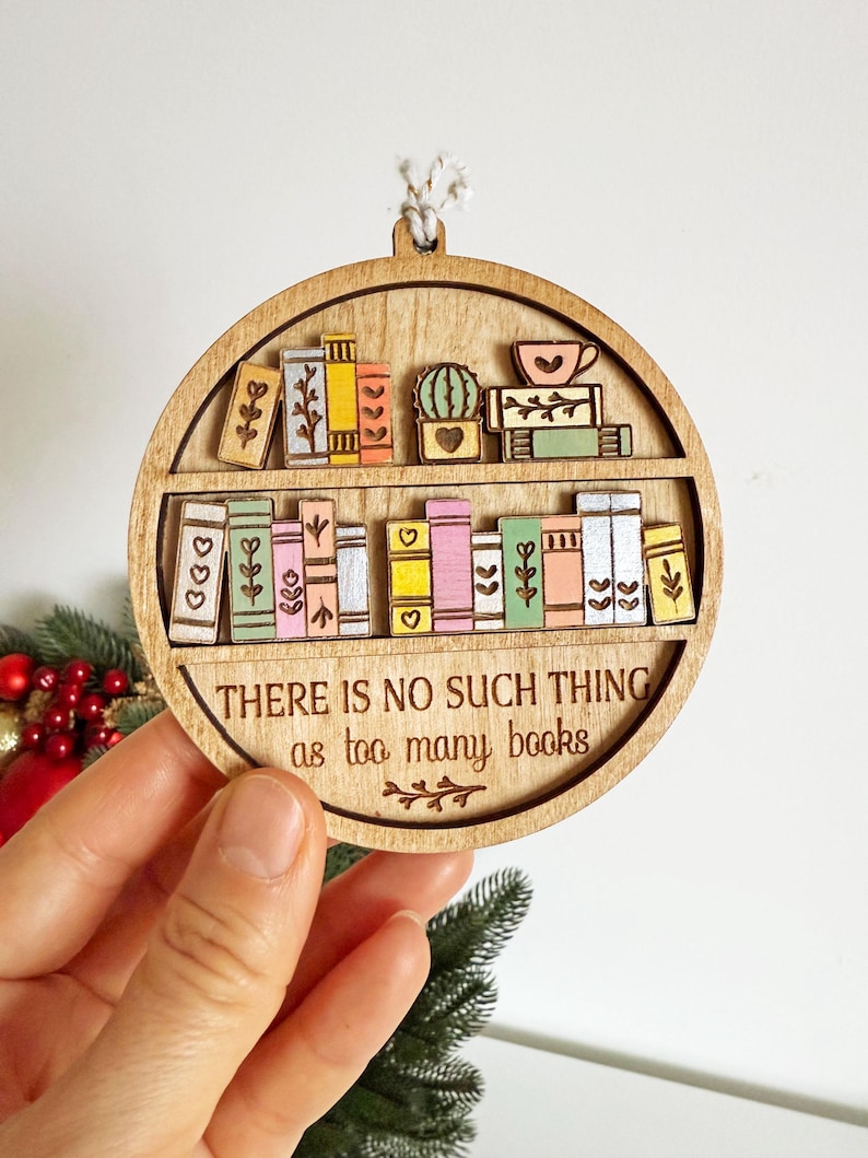 May include: Wooden, round ornament featuring a bookshelf design with colorful books and a cactus. The text "THERE IS NO SUCH THING as too many books" is engraved below the shelf. A decorative item for book lovers.
