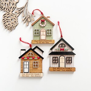 May include: Three wooden house-shaped Christmas ornaments with red hanging strings. One is green with a wreath, another is brown with a garland, and the third is white with a garland. Each has text: "Our First Christmas In Our New Home" or "THE WILSON FAMILY New Home New Memories."