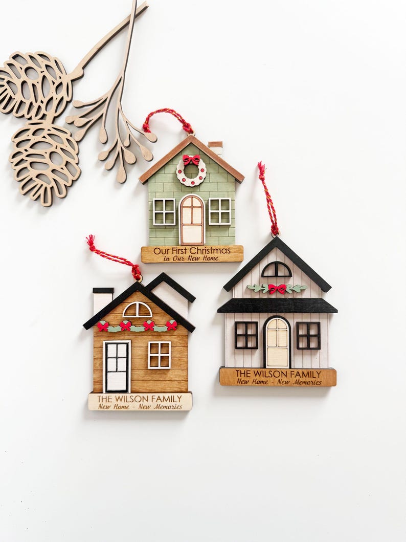 May include: Three wooden house-shaped Christmas ornaments with red hanging strings. One is green with a wreath, another is brown with a garland, and the third is white with a garland. Each has text: "Our First Christmas In Our New Home" or "THE WILSON FAMILY New Home New Memories."