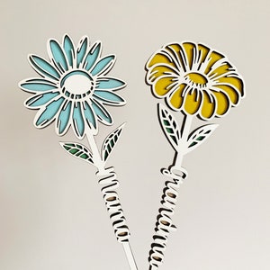 May include: Two wooden flower-shaped ornaments with the words "Grandma" and "Mama" written on the stems. The flowers are blue and yellow.