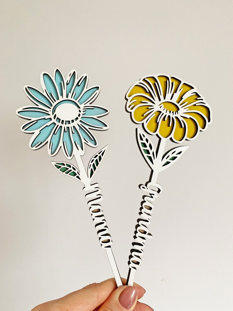 May include: Two wooden flower-shaped ornaments with the words "Grandma" and "Mama" written on the stems. The flowers are blue and yellow.