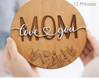 May include: A round wooden sign with the word "MOM" cut out, and the words "love you" in a cursive font. The sign has a floral design at the bottom and the text "2 Designs 12 Phrases". The sign is held by a person.