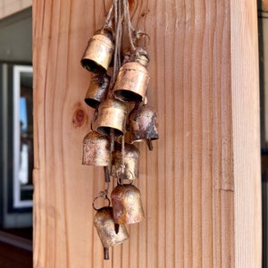 May include: A collection of small, gold-toned bells hanging from a wooden surface. The bells are tied together with twine and have a rustic, vintage look.