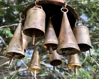 May include: A weathered metal wind chime featuring a copper-toned conical top and several bells of varying shapes. Some bells are cylindrical, others conical, all suspended by metal rings. The chime hangs amongst green tree branches.