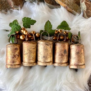 Peut inclure: Six cloches en métal de couleur dorée avec une finition vieillie. Les cloches sont disposées en rangée sur une surface blanche et moelleuse.