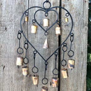 May include: A black metal heart-shaped wind chime with multiple small, aged brass bells. The bells hang from the heart's frame and are of varying sizes and shapes. The background is a weathered wooden surface.