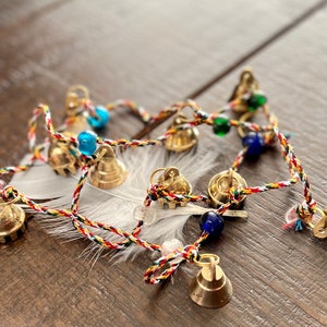 May include: A string of small gold bells with blue and green beads, tied together with a red, white, and yellow cord. The bells are hanging from a white feather.