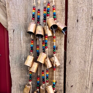 May include: A string of bells with colorful beads hanging from a wooden surface. The bells are gold and have a rustic look. The beads are a mix of red, orange, yellow, green, blue, and purple.