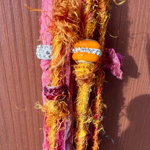 May include: Close-up of three strands of colorful hair extensions with beads and charms. The strands are a mix of orange, yellow, and pink. The beads are orange and clear with rhinestones. The charms are silver with rhinestones.