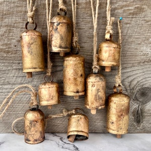 May include: A collection of nine rustic, gold-colored metal bells of varying sizes, suspended by natural twine. The bells have a distressed finish, and some show signs of wear. The background is a weathered wooden surface.