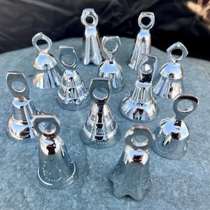 May include: A collection of 13 silver bells of various sizes and shapes. The bells have a shiny, polished finish and are arranged on a gray surface.
