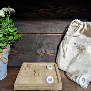 May include: A ceramic tic-tac-toe game set with white ceramic X's and O's. The game board is beige and has a rustic look. The game pieces are in a white drawstring bag with the text "Crescent Stone Pottery" printed on it.
