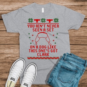 May include: A gray t-shirt with a Christmas-themed design featuring a red and green pattern and the text "You ain't never seen a set on a dog like this one's got Clark".