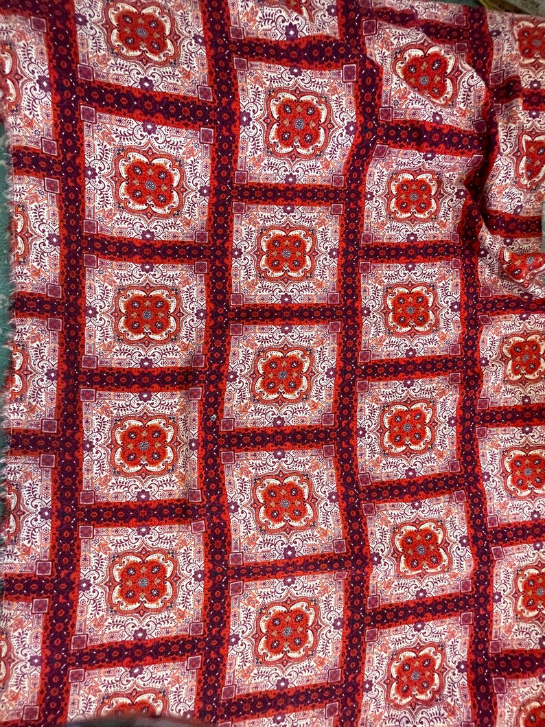 K&ouml;nnte beinhalten: Roter und wei&szlig;er Stoff mit einem sich wiederholenden Muster aus komplizierten quadratischen Designs. Der Stoff hat ein paisleyartiges Design mit einem burgunderfarbenen Hintergrund.