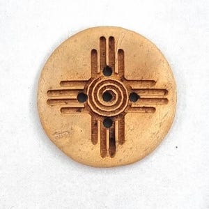 May include: A round wooden ornament with a carved design of the Zia symbol, a traditional symbol of the Pueblo people of New Mexico. The symbol is a four-pointed star with a circle in the center.