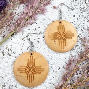 May include: Two round, light brown earrings with a carved sun symbol design. The earrings have a spiral center and straight lines radiating outward. They hang from silver-colored fish hook ear wires. The background includes dried purple flowers.