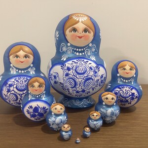 Pu&ograve; includere: Un set di dieci bambole di legno nidificate con un motivo floreale blu e bianco. Ogni bambola ha un volto dipinto con capelli castani, una collana di perle bianche e intricati disegni a volute blu sulla parte inferiore del corpo.