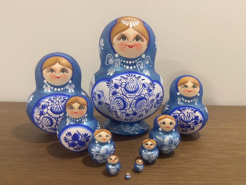 Pu&ograve; includere: Un set di dieci bambole di legno nidificate con un motivo floreale blu e bianco. Ogni bambola ha un volto dipinto con capelli castani, una collana di perle bianche e intricati disegni a volute blu sulla parte inferiore del corpo.