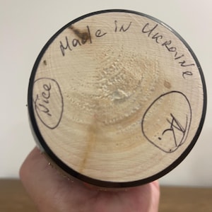 K&ouml;nnte beinhalten: Ein Holzobjekt mit kreisf&ouml;rmiger Basis, mit dem handgeschriebenen Text "Made in Ukraine". Die Basis ist hellbraun mit dunklem Rand. Das Wort "Nice" ist in einem Kreis geschrieben. Das Objekt wird in einer Hand gehalten.
