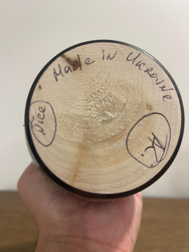 K&ouml;nnte beinhalten: Ein Holzobjekt mit kreisf&ouml;rmiger Basis, mit dem handgeschriebenen Text "Made in Ukraine". Die Basis ist hellbraun mit dunklem Rand. Das Wort "Nice" ist in einem Kreis geschrieben. Das Objekt wird in einer Hand gehalten.