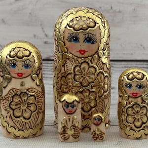 May include: A set of five Russian nesting dolls, or Matryoshka dolls, in varying sizes. Each doll features a cream-colored base with gold accents, including floral patterns and facial details. The dolls have blue eyes and red lips.