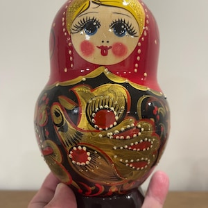 K&ouml;nnte beinhalten: Traditionelle russische Matrjoschka-Puppe mit rotem Kopftuch und bemaltem Gesicht. Der K&ouml;rper der Puppe ist mit schwarzen und goldenen Blumenmustern und roten Akzenten verziert. Der Sockel ist dunkelbraun.