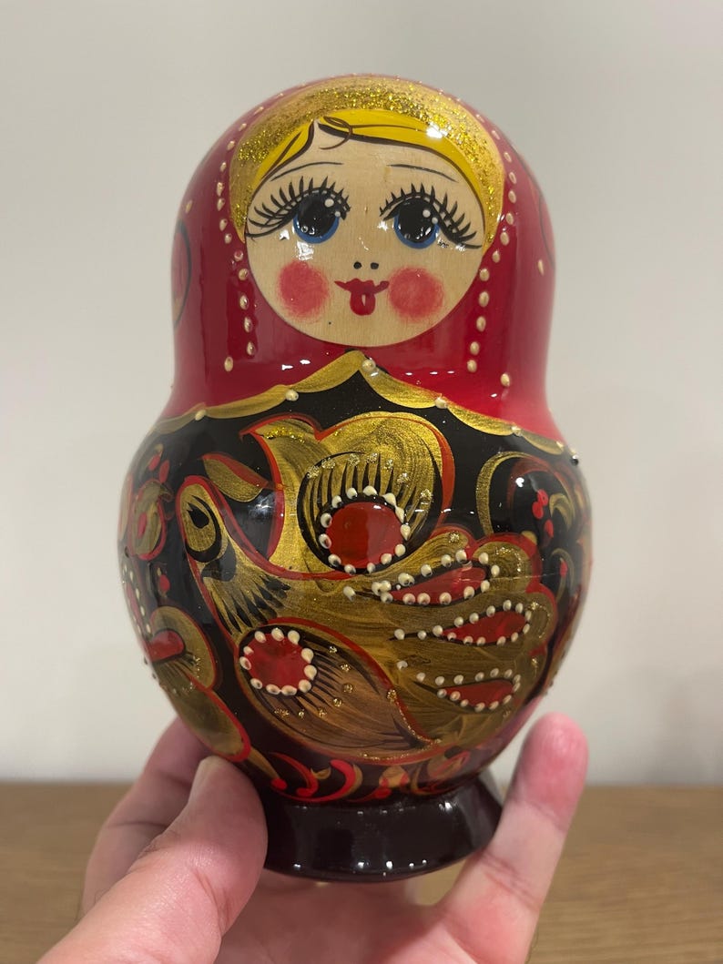 K&ouml;nnte beinhalten: Traditionelle russische Matrjoschka-Puppe mit rotem Kopftuch und bemaltem Gesicht. Der K&ouml;rper der Puppe ist mit schwarzen und goldenen Blumenmustern und roten Akzenten verziert. Der Sockel ist dunkelbraun.