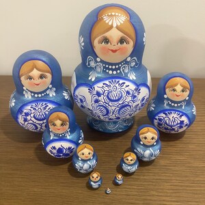 Pu&ograve; includere: Un set di dieci bambole in legno nidificate con una combinazione di colori blu e bianco. Ogni bambola &egrave; dipinta a mano con un viso sorridente, capelli castano chiaro e intricati motivi floreali blu sulla parte inferiore del corpo.