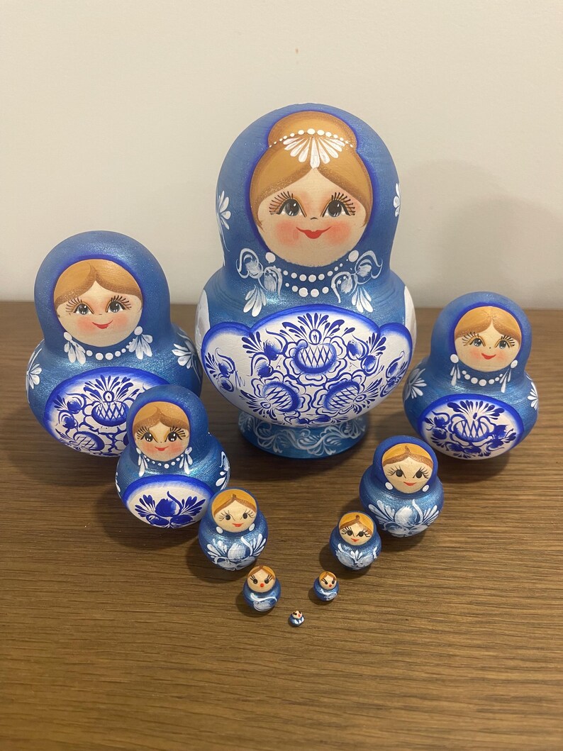 Pu&ograve; includere: Un set di dieci bambole in legno nidificate con una combinazione di colori blu e bianco. Ogni bambola &egrave; dipinta a mano con un viso sorridente, capelli castano chiaro e intricati motivi floreali blu sulla parte inferiore del corpo.