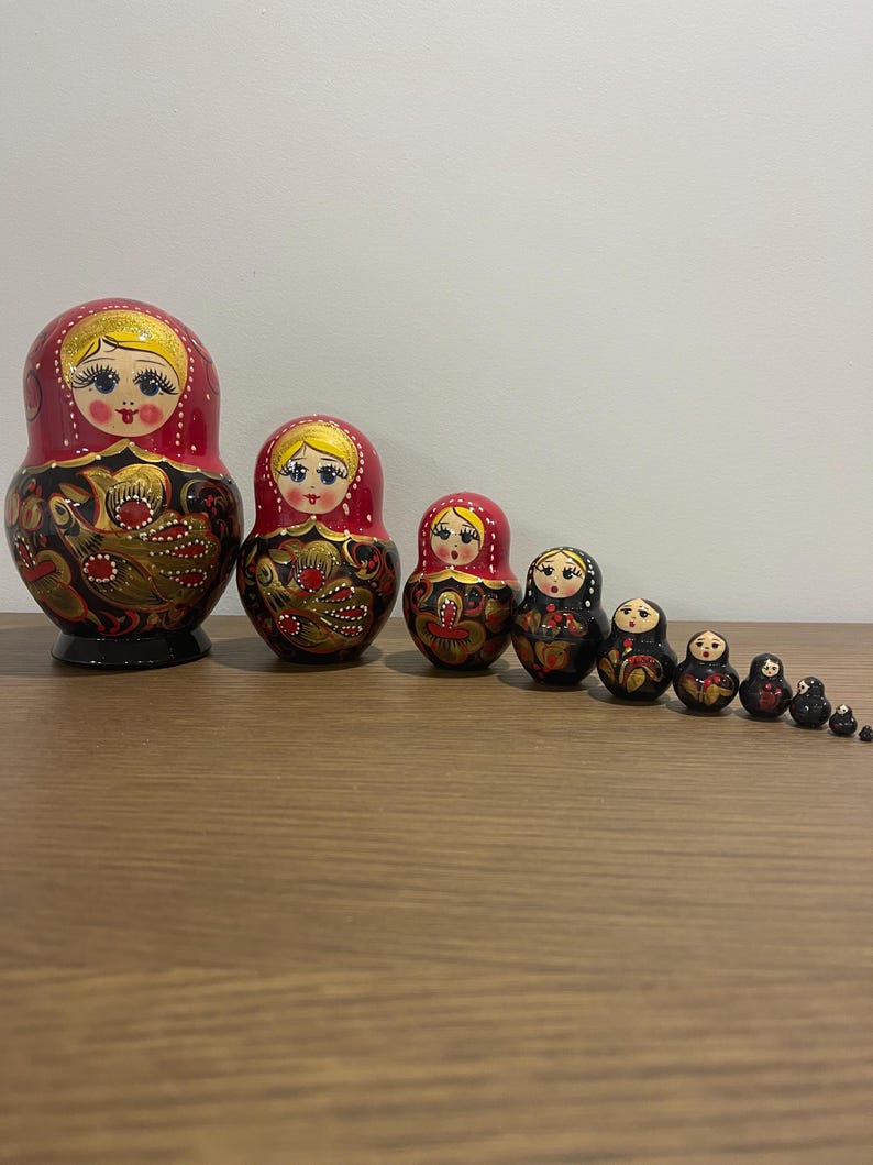 K&ouml;nnte beinhalten: Eine Reihe traditioneller russischer Matrjoschka-Puppen. Die Puppen sind in Rot, Schwarz und Gold bemalt und weisen Blumen- und Vogelmuster auf. Die gr&ouml;&szlig;te Puppe ist etwa 15 cm hoch.