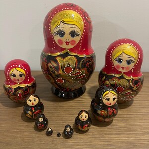 K&ouml;nnte beinhalten: Eine Sammlung russischer Matrjoschka-Puppen in verschiedenen Gr&ouml;&szlig;en. Die Puppen sind in Rot und Schwarz mit goldenen Akzenten bemalt. Die gr&ouml;&szlig;te Puppe ist etwa 18 cm hoch, die kleinste etwa 2,5 cm.