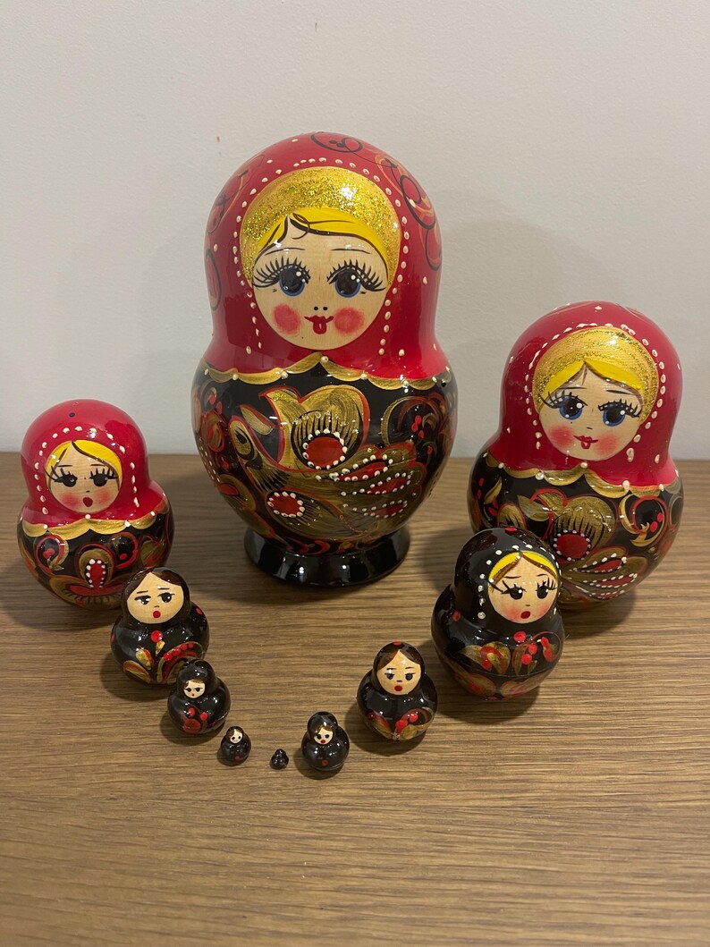 K&ouml;nnte beinhalten: Eine Sammlung russischer Matrjoschka-Puppen in verschiedenen Gr&ouml;&szlig;en. Die Puppen sind in Rot und Schwarz mit goldenen Akzenten bemalt. Die gr&ouml;&szlig;te Puppe ist etwa 18 cm hoch, die kleinste etwa 2,5 cm.