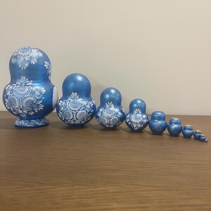 Pu&ograve; includere: Un set di dieci bambole matrioska disposte in fila dalla pi&ugrave; grande alla pi&ugrave; piccola. Ogni bambola presenta una finitura blu metallizzata con intricati motivi floreali bianchi dipinti sul davanti, terminando con un minuscolo pezzo blu.