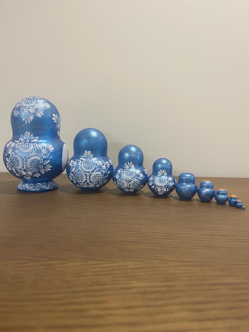 Pu&ograve; includere: Un set di dieci bambole matrioska disposte in fila dalla pi&ugrave; grande alla pi&ugrave; piccola. Ogni bambola presenta una finitura blu metallizzata con intricati motivi floreali bianchi dipinti sul davanti, terminando con un minuscolo pezzo blu.
