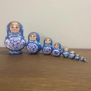 Pu&ograve; includere: Un set di dieci matrioske blu e bianche disposte in fila dalla pi&ugrave; grande alla pi&ugrave; piccola. Ogni bambola presenta un volto dipinto a mano con capelli castani e un dettagliato motivo floreale sul corpo, realizzato con vernice blu e bianca.