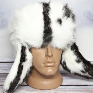 May include: A white fur hat with black stripes and spots, featuring ear flaps. The hat is displayed on a mannequin head against a winter-themed backdrop. The hat's design includes a mix of white and black fur.