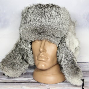 May include: A gray fur hat with ear flaps, displayed on a mannequin head. The hat has a fluffy texture and a neutral color palette. The ear flaps are designed to be tied under the chin. The background is a winter scene.