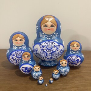 Pu&ograve; includere: Un set di dieci bambole matrioska con una tavolozza di colori blu e bianco. Ogni bambola presenta un volto dipinto a mano con capelli castano chiaro e intricati motivi floreali blu sulla parte inferiore del corpo.