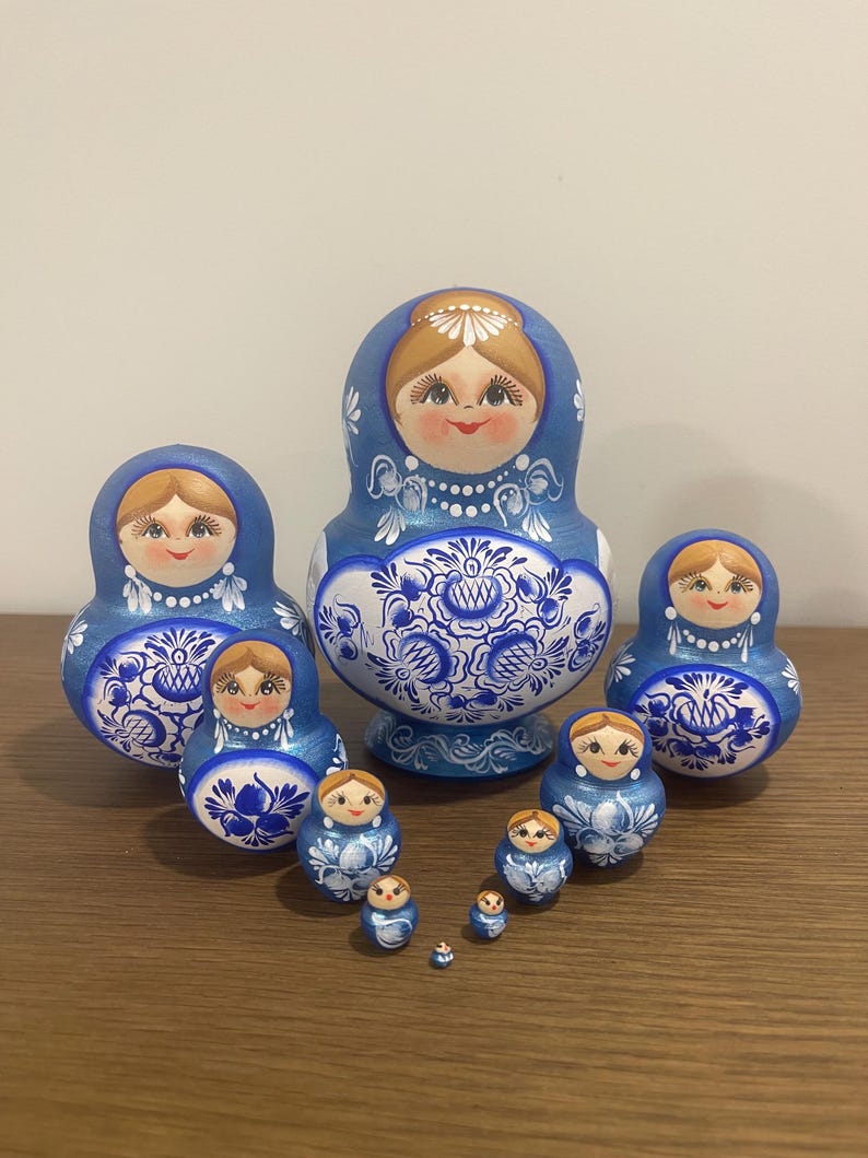 Pu&ograve; includere: Un set di dieci bambole matrioska con una tavolozza di colori blu e bianco. Ogni bambola presenta un volto dipinto a mano con capelli castano chiaro e intricati motivi floreali blu sulla parte inferiore del corpo.