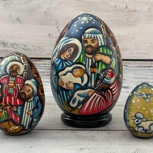 May include: Three hand-painted wooden Easter eggs with religious scenes. The largest egg depicts the Nativity, the middle egg shows the three wise men, and the smallest egg features a lamb and a dove. The eggs are on a light wood surface.