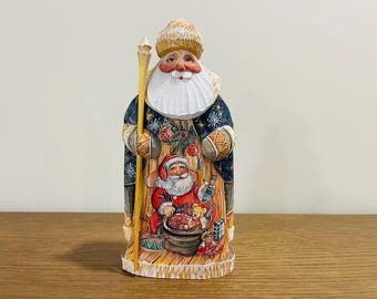 Op de afbeelding: Een handgesneden houten kerstmanfiguur met een donkerblauwe jas met sneeuwvlokpatronen, een witte baard en een lange gele staf. De voorkant van de jas toont een geschilderde scène van een kleinere kerstman met speelgoed en een zak met cadeaus.