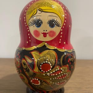 K&ouml;nnte beinhalten: Traditionelle russische Matrjoschka-Puppe mit rotem Kopf und K&ouml;rper. Die Puppe hat ein bemaltes Gesicht mit blauen Augen, rosigen Wangen und einem roten Mund. Der untere Teil ist schwarz mit goldenen und roten Blumenmustern.