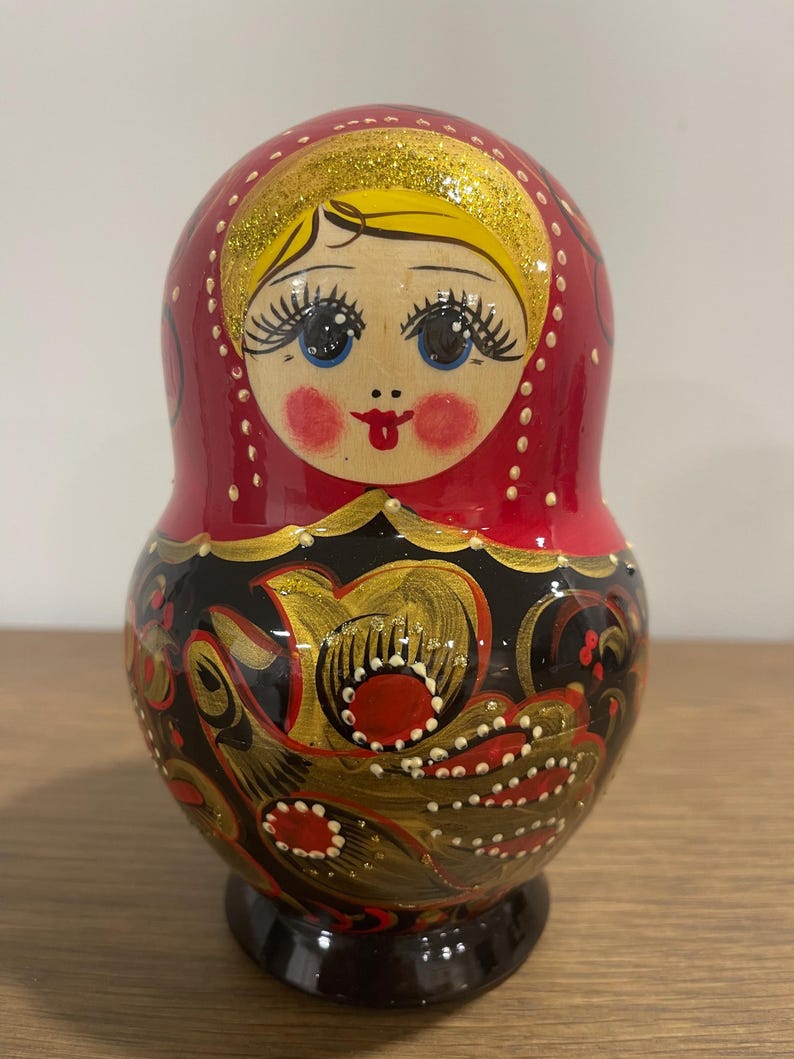 K&ouml;nnte beinhalten: Traditionelle russische Matrjoschka-Puppe mit rotem Kopf und K&ouml;rper. Die Puppe hat ein bemaltes Gesicht mit blauen Augen, rosigen Wangen und einem roten Mund. Der untere Teil ist schwarz mit goldenen und roten Blumenmustern.