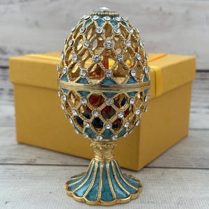 May include: An ornate, egg-shaped metal trinket box featuring a gold-tone lattice design with teal enamel accents and embedded clear rhinestones. The piece stands on a flared, fluted base and is topped with a single, larger clear crystal.