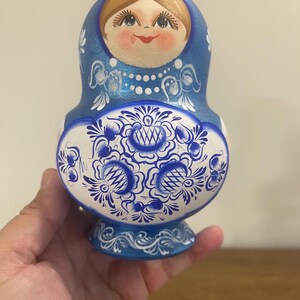 Pu&ograve; includere: Una matrioska in legno blu e bianca con un volto dipinto a mano, capelli castani e un motivo floreale dettagliato sulla parte inferiore. Il design include accenti decorativi bianchi, una collana di perle e una base blu solida.