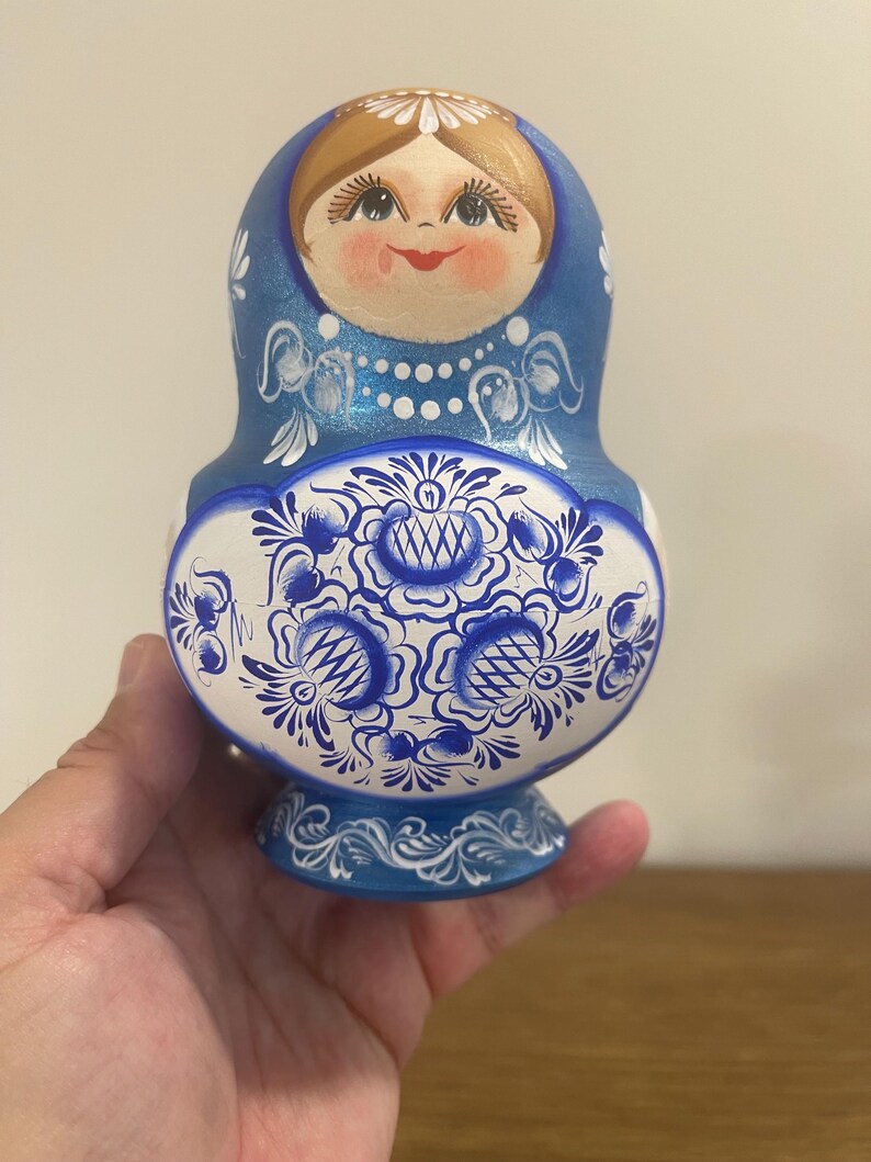 Pu&ograve; includere: Una matrioska in legno blu e bianca con un volto dipinto a mano, capelli castani e un motivo floreale dettagliato sulla parte inferiore. Il design include accenti decorativi bianchi, una collana di perle e una base blu solida.