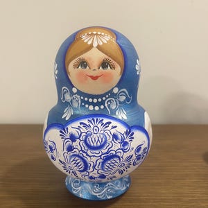 Pu&ograve; includere: Una matrioska in legno blu con un volto dipinto a mano, capelli castani, un copricapo floreale bianco e labbra rosse. La sezione inferiore presenta intricati motivi floreali blu e bianchi con delicati ghirigori e punti bianchi.
